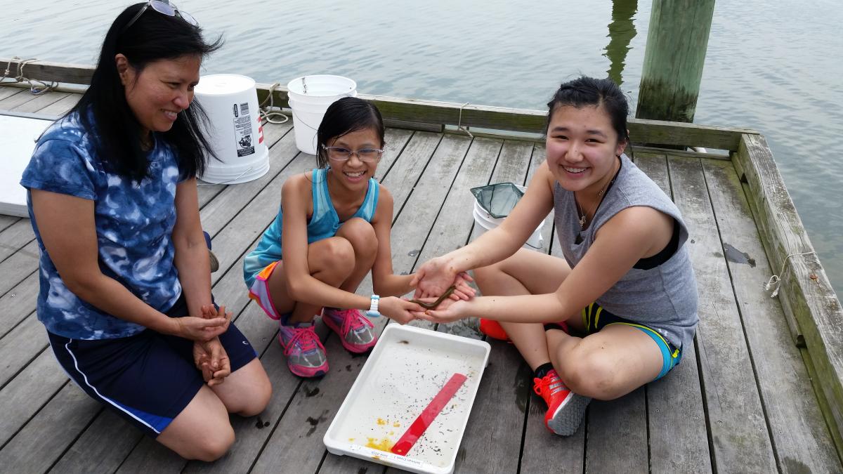 Three people smiling and posing with an eel over a sampling container on a wooden dock against a backdrop of water.