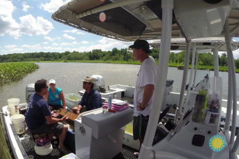 2 men & 1 woman sitting and talking on a boat. Another man drives the boat through the Patuxent River, under partly cloudy skies