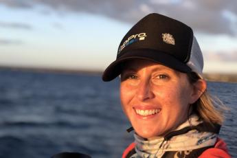 Head and shoulders photo of a young woman wearing a scarf and a black and gray cap, with the ocean behind her