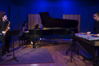 A man playing saxophone, a woman playing piano, and a man playing percussion in a room with blue backlighting