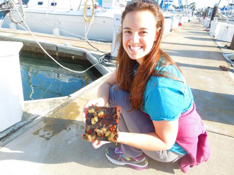 A white woman smiles at the camera while holding a sample device on a bright sunny doc. 