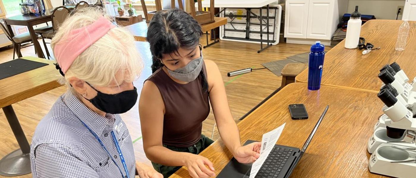 Two women at a laptop entering data from cellphone app. 