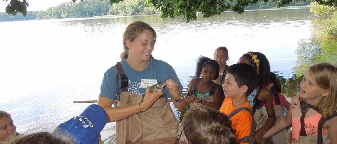 Young woman in waders stands on a beach, holding a fish, as a group of schoolchildren also in waders look on