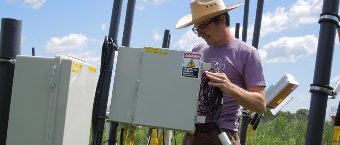 Man in straw hat beside gray electrical box on grassy marsh