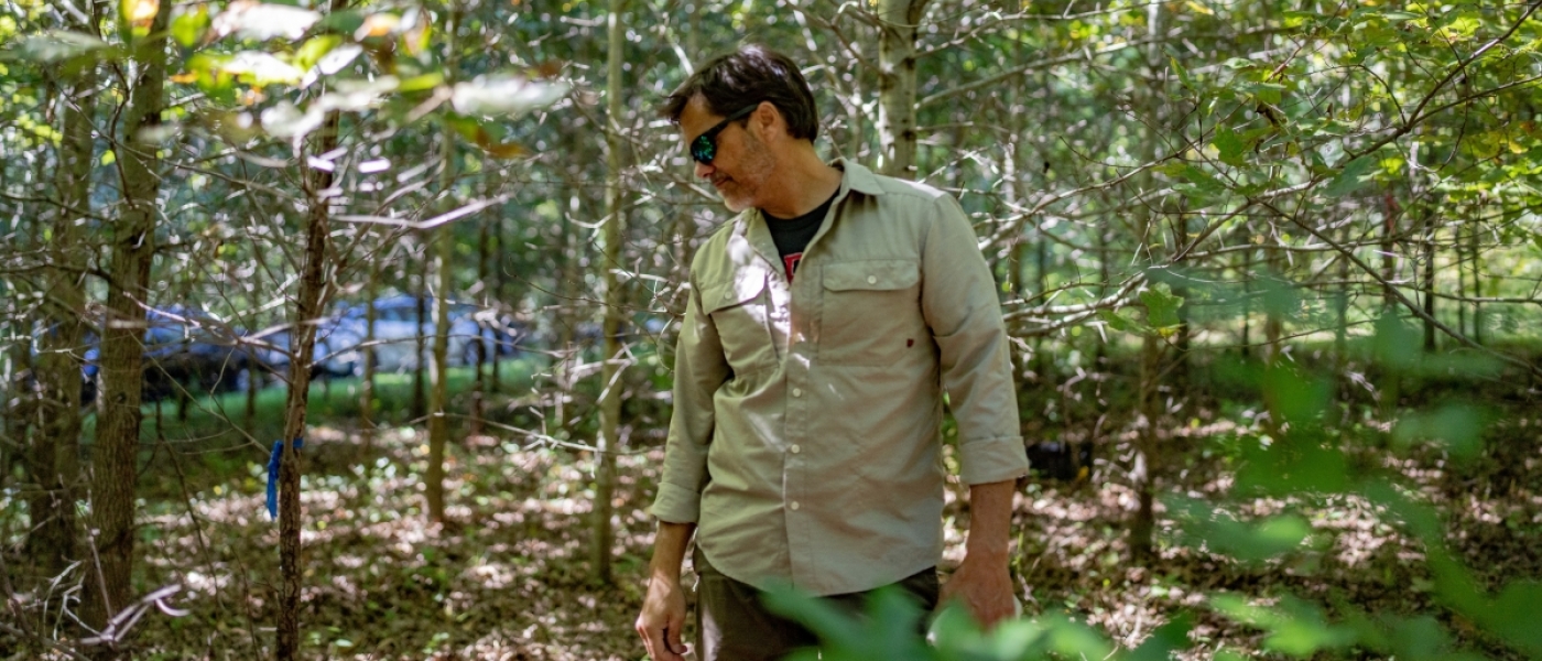 A man in a tan colored shirt and sunglasses poses, looking to his left and down, in the middle of a forest