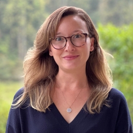 A headshot of Anna Davis, who has shoulder-length blond hair, glasses, and is wearing a blue sweater, in front of a forest.