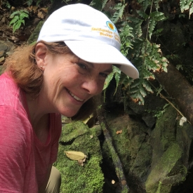 Photo of Cole Johnson, with short hair, wearing a red shirt and white cap, smiling in front of green leaves