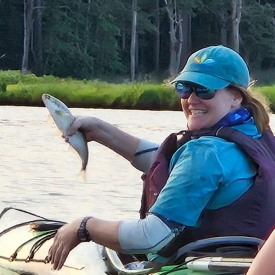 Ray, wearing a hat and sunglasses, sits in a kayak on the water holding a medium sized fish. She looks excited.