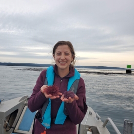 photo of Mary on a boat holding a red sea urchin in one hand and a purple sea urchin in the other